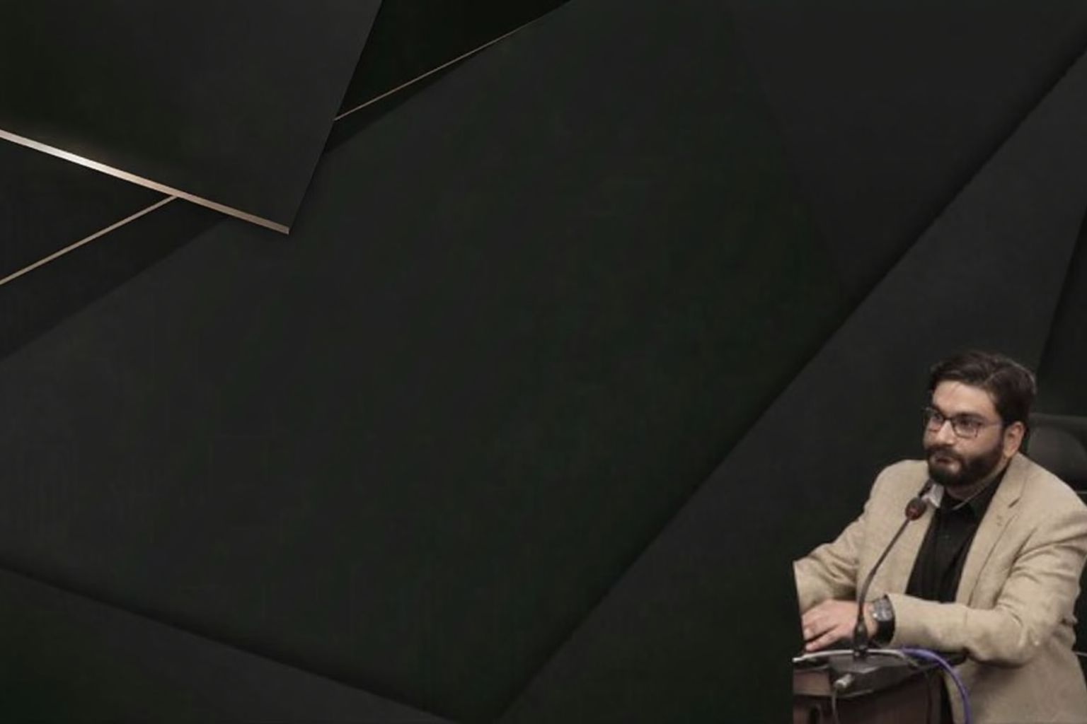 Mand med skæg og briller sidder ved et bord med mikrofon, delvist skjult af mørke former.